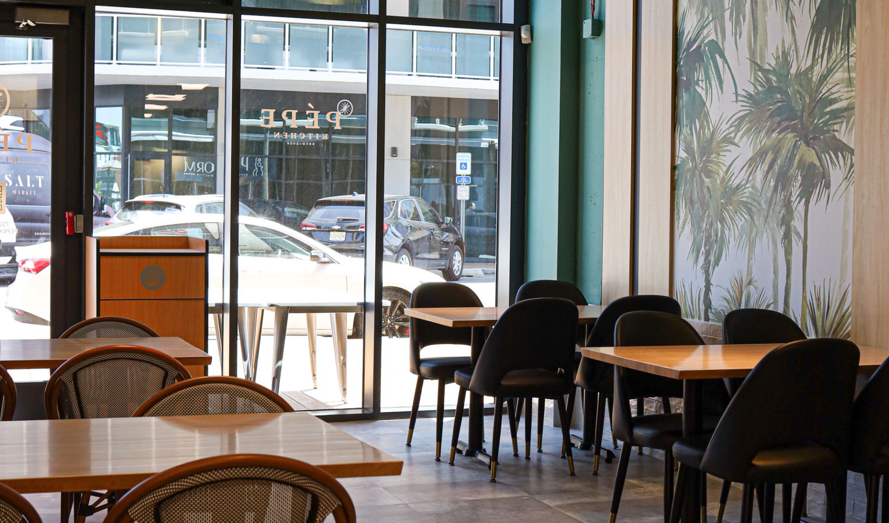 Window seating at Pēpe Kitchen with oak tables, bistro chairs, and palm-print wallpaper